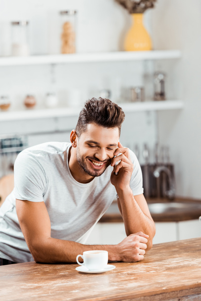smiling young man talking by smartphone while leaning at kitchen table with cup of coffee - Foto, imagen
