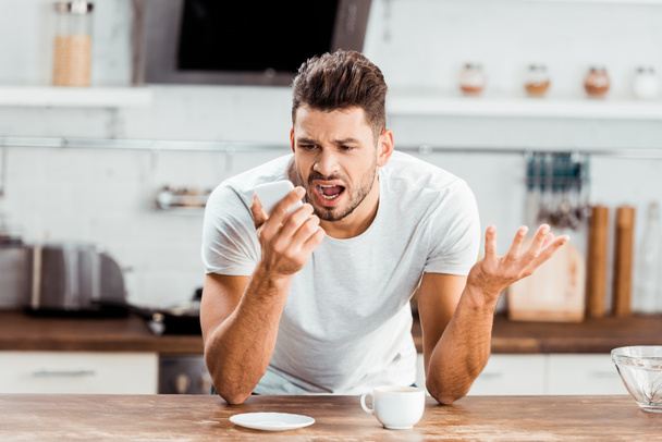emotional young man yelling at smartphone while drinking coffee in kitchen - Photo, Image