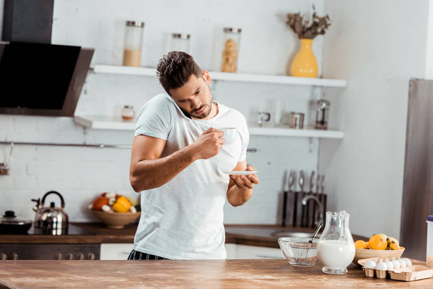 young man holding cup of coffee and talking by smartphone in kitchen - Foto, Bild