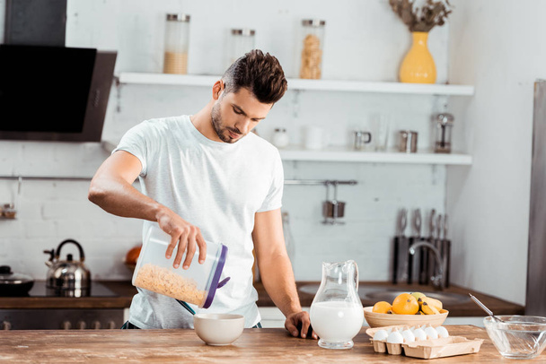 young man holding container with corn flakes and preparing breakfast in kitchen - Foto, Imagen