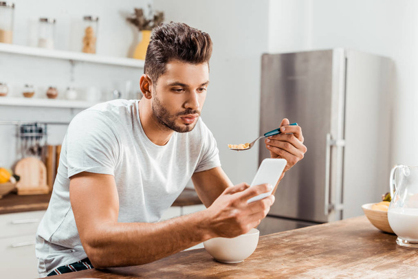 handsome young man using smartphone and having breakfast at home - Foto, Imagen