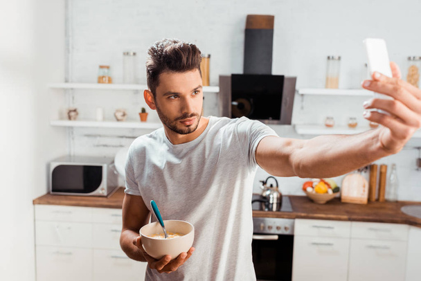 przystojny, młody człowiek gospodarstwa miskę z śniadanie i biorąc selfie z smartphone w kuchni - Zdjęcie, obraz