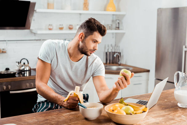 young man in pajamas holding fruits and using laptop at morning - Foto, immagini