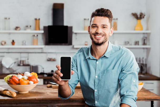 handsome young man holding smartphone with blank screen and smiling at camera in kitchen - Valokuva, kuva
