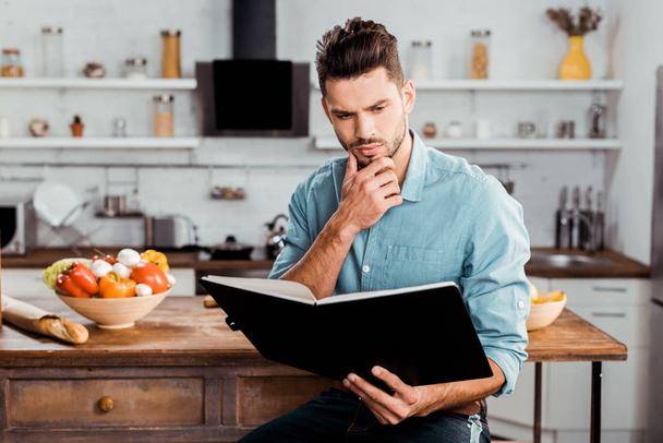 thoughtful handsome young man reading cookbook in kitchen - Foto, Bild