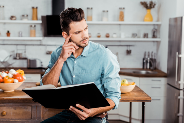 pensive young man holding cookbook and looking away in kitchen - Foto, Bild