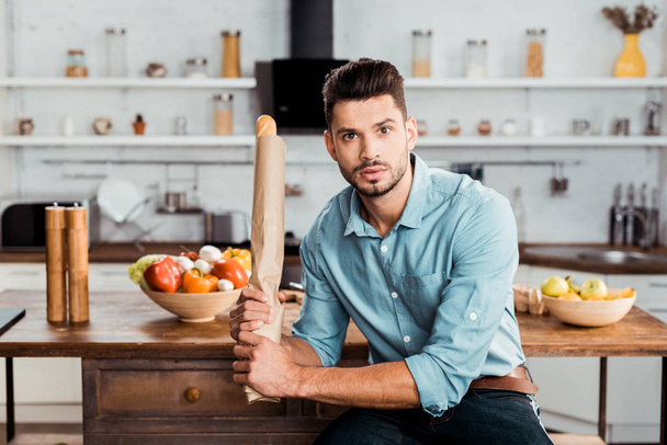 handsome young man holding fresh baguette in paper bag and looking at camera in kitchen - Foto, Bild