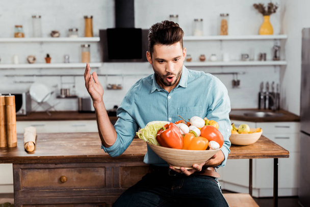 возбужденный молодой человек держит миску со свежими овощами на кухне
 - Фото, изображение