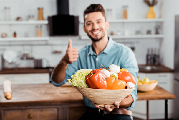 Lächelnder junger Mann hält Schüssel mit frischem Gemüse in der Hand und zeigt Daumen hoch in der Küche  - Foto, Bild