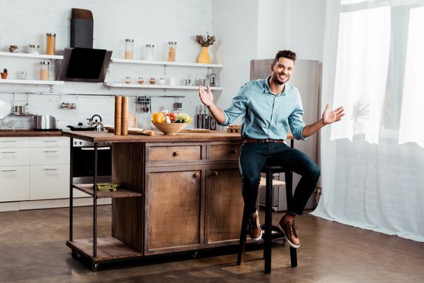 cheerful young man smiling at camera while sitting with open arms in kitchen - Foto, Imagem