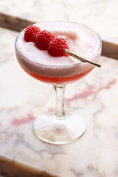 Pink Clover Club Cocktail Drink in Coupe Glass with Layer of Foam and Raspberry Garnish on Minimalist Marble Background - Фото, изображение
