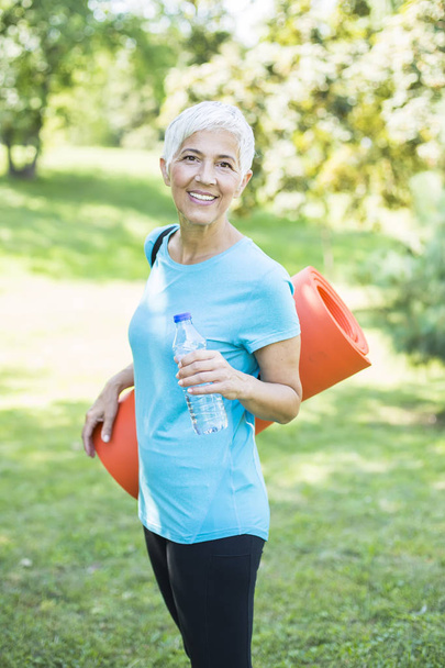 Retrato de mulher sênior mantém tapete de fitness em suas costas no parque e se preparando para o exercício
 - Foto, Imagem