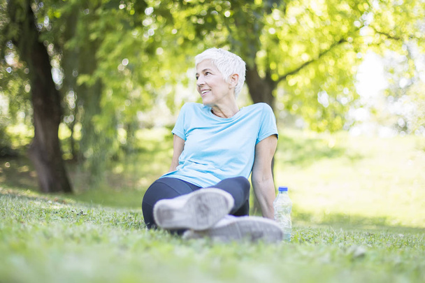 Vanhempi nainen istuu ja lepää harjoituksen jälkeen puistossa nurmella
 - Valokuva, kuva