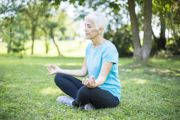 Senior woman in a lotus position   on a grass in park - Photo, Image