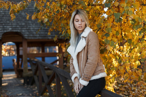 Fashionable attractive young blonde girl in a stylish autumn beige jacket and black trousers sitting on the bridge on the background of a tree. Autumn, a woman on a background of golden autumn leaves - Photo, Image