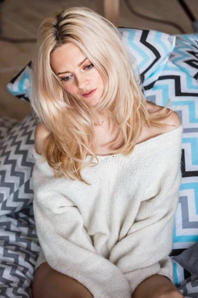 Studio portrait of beauty blond woman wearing white sweater. Female model with stylish makeup lying in bed - Photo, image