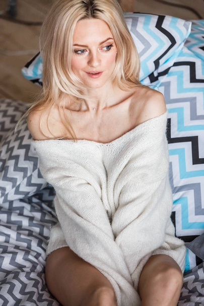 Studio portrait of beauty blond woman wearing white sweater. Female model with stylish makeup lying in bed - Foto, imagen
