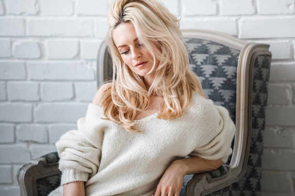 Studioporträt einer schönen blonden Frau mit weißem Pullover. weibliches Modell mit stilvollem Make-up sitzt im Sessel, Backsteinwand Hintergrund - Foto, Bild