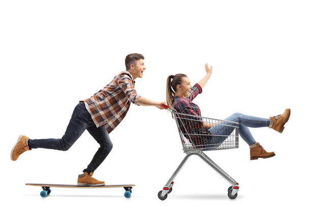Full length shot of a young guy riding a longboard and pushing a girl in a shopping cart isolated on white background - Foto, Bild