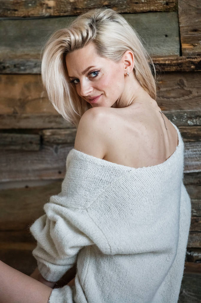 Studio portrait of beauty blond woman wearing white sweater. Female model sitting on wooden floor  - Photo, image