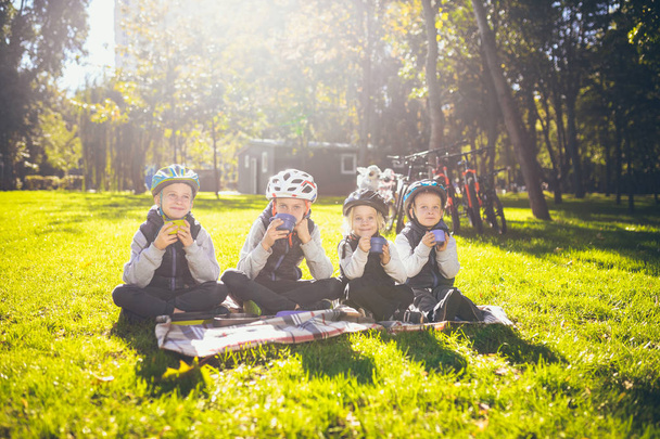 Tema activo vacaciones en familia naturaleza. grupo de personas niños pequeños tres hermanos y hermanas se sientan en la manta cerca de las bicicletas en el parque verde césped césped descanso y beber bebida de tazas y termos té
. - Foto, imagen
