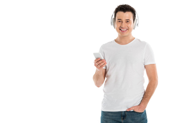 hombre guapo en auriculares con teléfono inteligente y mirando a la cámara aislada en blanco
 - Foto, Imagen