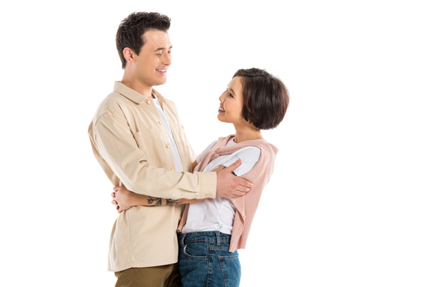 Glimlachende man en mooie vrouw in casual kleding knuffelen en kijken elkaar geïsoleerd op wit - Foto, afbeelding