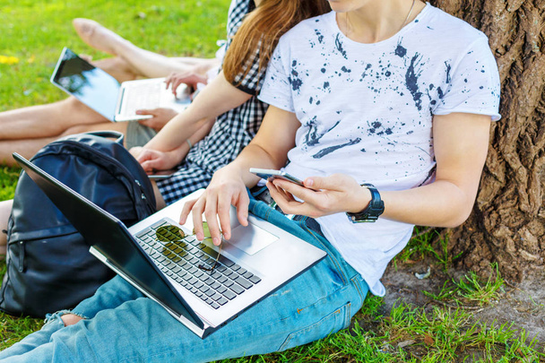 Distance learning concept. Group of students in a park with open laptops sitting on the grass under a tree - Foto, afbeelding