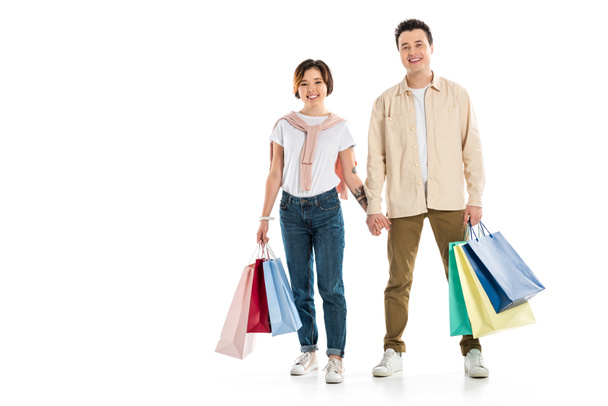 smiling couple looking at camera, holding hands and carrying shopping bags isolated on white  - 写真・画像