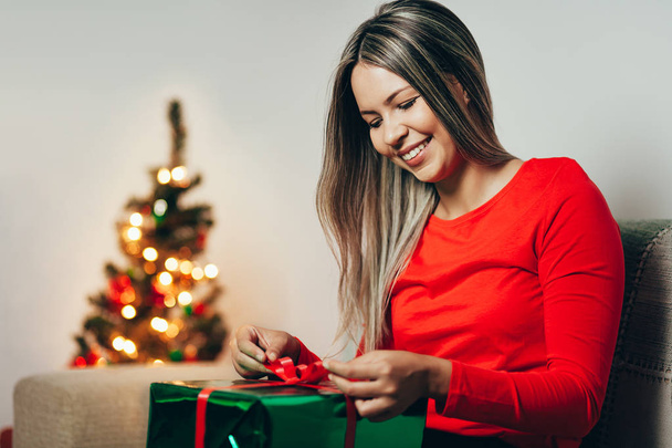 Young woman opening Christmas gift, sitting in the living room - Valokuva, kuva