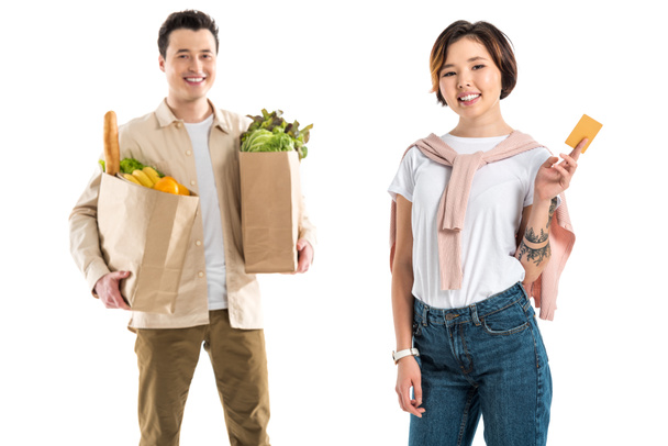 smiling husband holding grocery bag while wife presenting credit card isolated on white - Foto, afbeelding