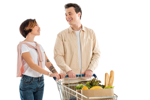 smiling young couple with shopping cart full of groceries looking at each other isolated on white - Foto, Imagem