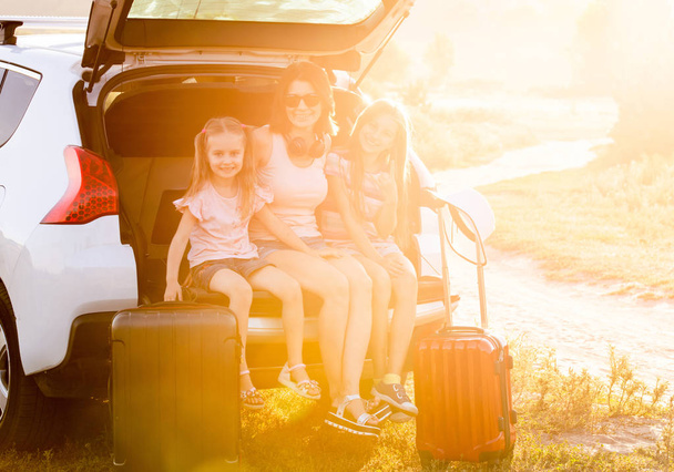 Мама и маленькие дочери сидят, обнимаясь на багажнике машины во время отпуска
 - Фото, изображение