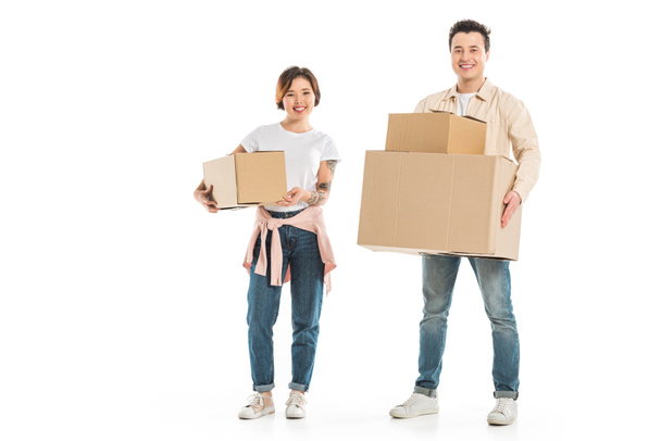 happy couple looking at camera and holding cardboard boxes isolated on white, moving to new house concept - Фото, изображение