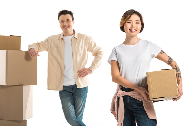 smiling couple looking at camera and holding cardboard boxes isolated on white, moving to new house concept - Foto, immagini