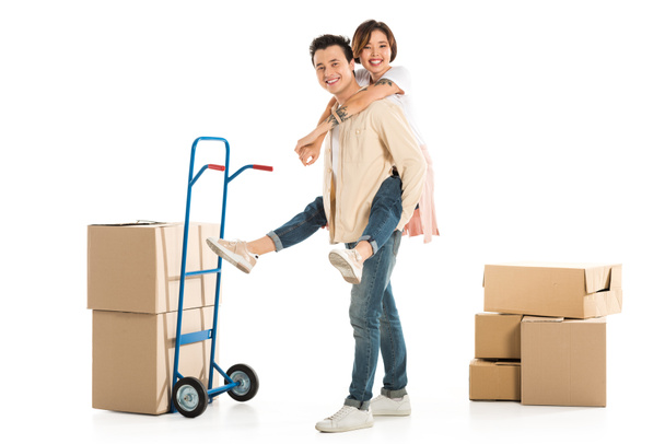 husband giving piggyback ride to wife near cardboard boxes and hand truck on background, moving to new house concept - Photo, Image
