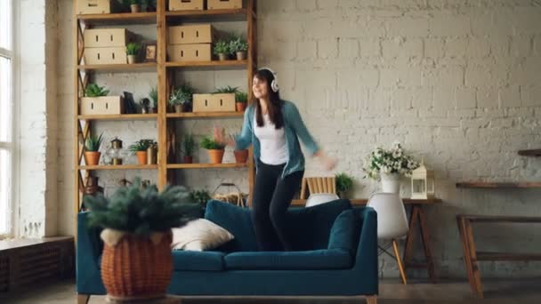 Funny woman is dancing and jumping on sofa listening to music through headphones having fun and enjoying freedom and leisure. Loft style apartment is in background. - Filmati, video