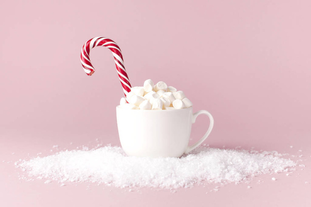 Christmas New Year drink, white mug with marshmallows and Candy Cane in the snow on pink background Flat Lay copy space. Winter traditional drink food. Festive decor celebration Xmas holiday 2019 - Photo, Image