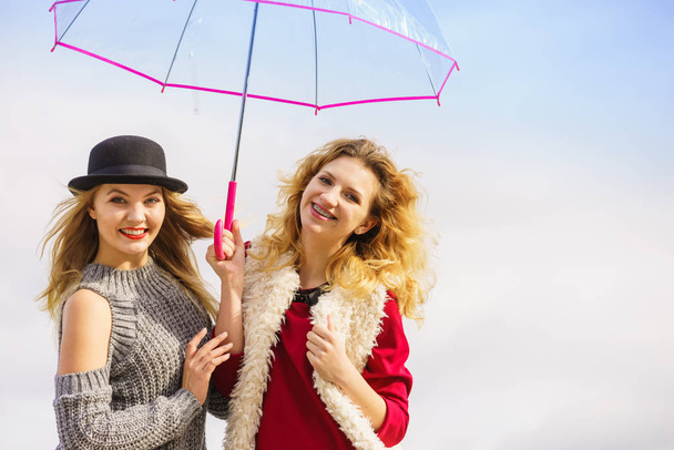 Two fashionable women wearing stylish outfits holding transparent umbrella spending their free time outdoor - Foto, Imagem