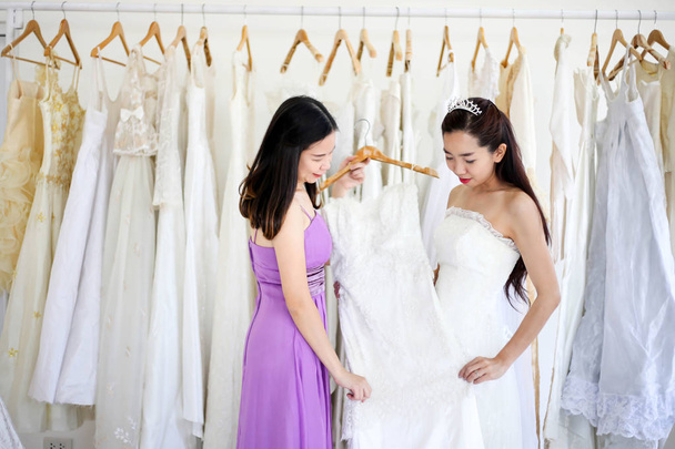 Beautiful bride getting dressed by her best friend in her wedding day and choosing a wedding dress in the shop and the shop assistant is helping her - Photo, image