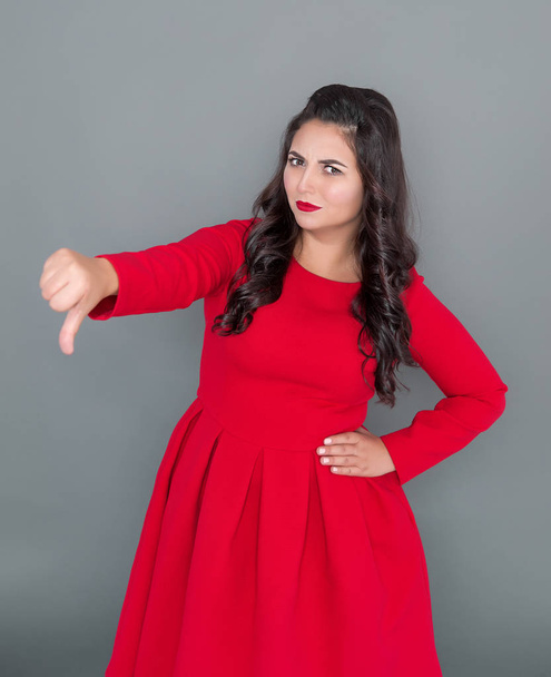 Плюс размер женщины в красном платье палец вниз жест на сером фоне
 - Фото, изображение