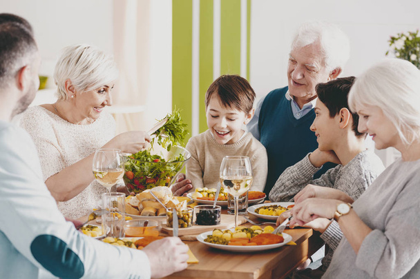 Hymyilevä poika istuu isoisän polvillaan perheen illallisen aikana vanhempien ja isoäidin kanssa.
 - Valokuva, kuva