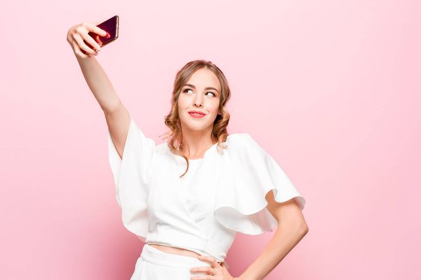 Portrait of a young attractive woman making selfie photo with smartphone on a pink background - 写真・画像