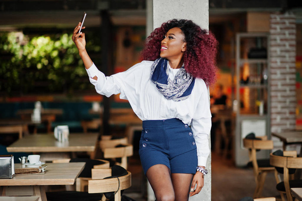 Attractive african american curly girl with mobile phone at hand posed at cafe and making selfie. - Foto, Imagem