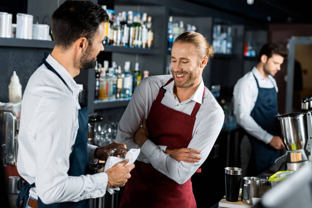 веселые улыбающиеся бармены, стоящие со скрещенными руками на рабочем месте, в то время как коллега полирует стекло
 - Фото, изображение