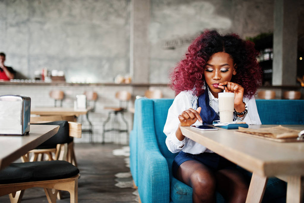 Attractive african american curly girl sitting at cafe drinking latte and looking at mobile phone. - Foto, Bild