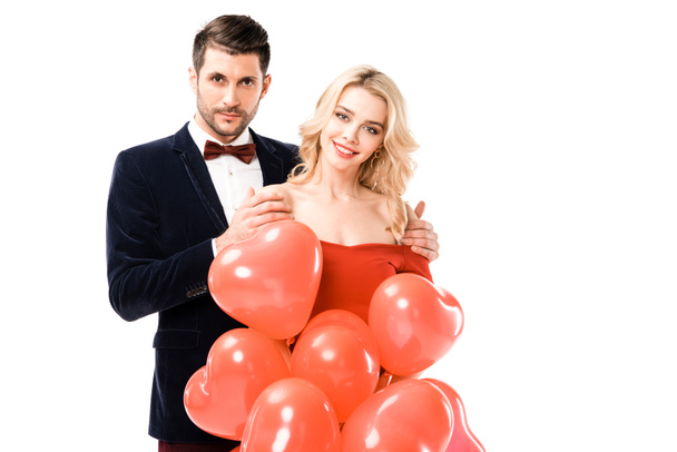 Ευτυχισμένο ζευγάρι βλέπει την κάμερα ενώ κρατάτε κόκκινα μπαλόνια που απομονώνονται σε λευκό - Φωτογραφία, εικόνα