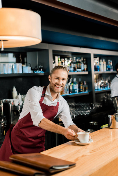 adult smiling barista in apron serving coffee at wooden counter - Φωτογραφία, εικόνα