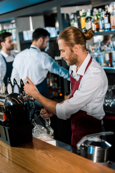 barman in apron pouring beer in glass behind wooden counter at bar - Photo, Image