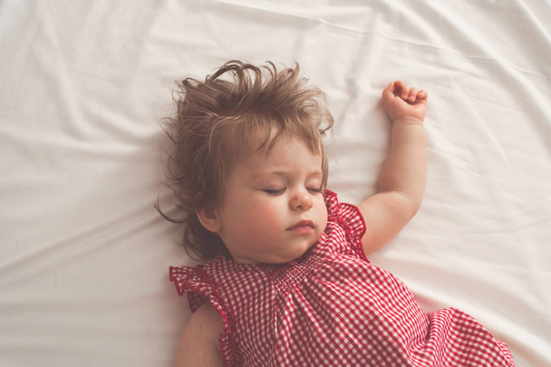 Κοριτσάκι κοιμάται ανάσκελα με ανοιχτές αγκάλες και χωρίς πιπίλα σε ένα κρεβάτι με λευκά σεντόνια. Ήσυχο ύπνο σε ένα φωτεινό δωμάτιο. Ρετρό παστέλ τονισμένα. Απαλή εστίαση - Φωτογραφία, εικόνα